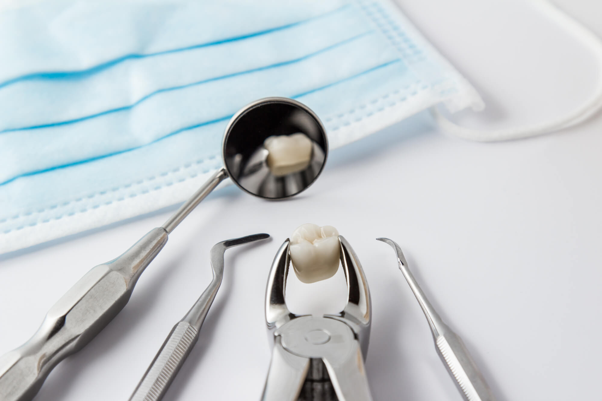 Tooth extraction concept with an array of stainless steel dental tools and a mask with the extracted tooth clasped in the pincers and reflected in the mirror